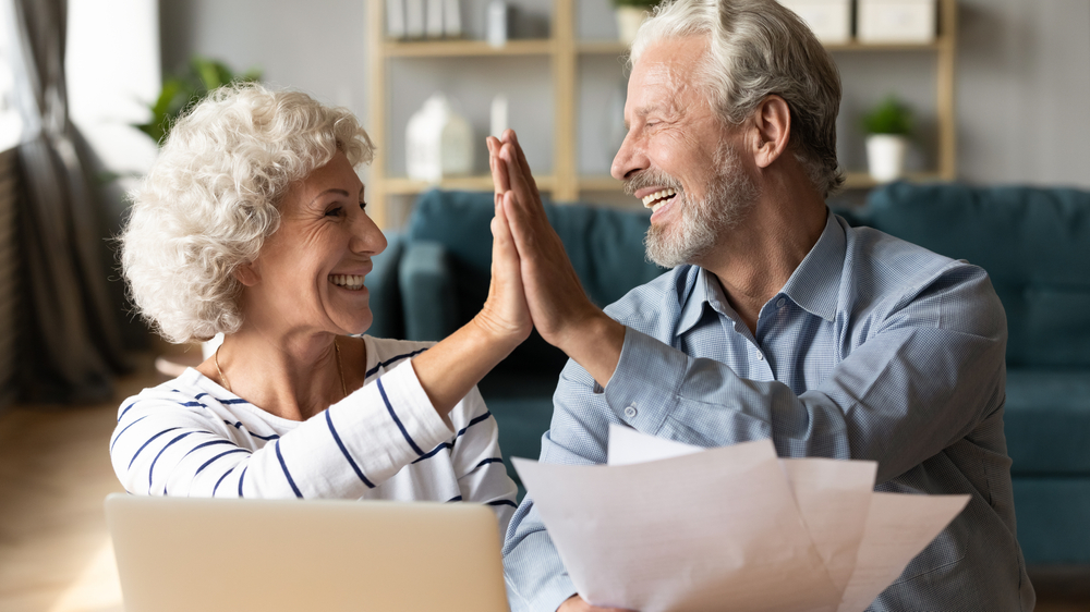 A senior woman high fives her husband after looking over a laptop and paperwork