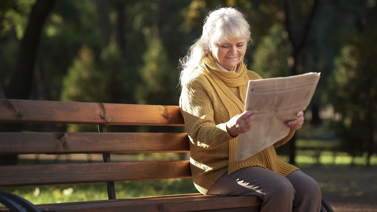 senior woman reading a newspaper on a park bench